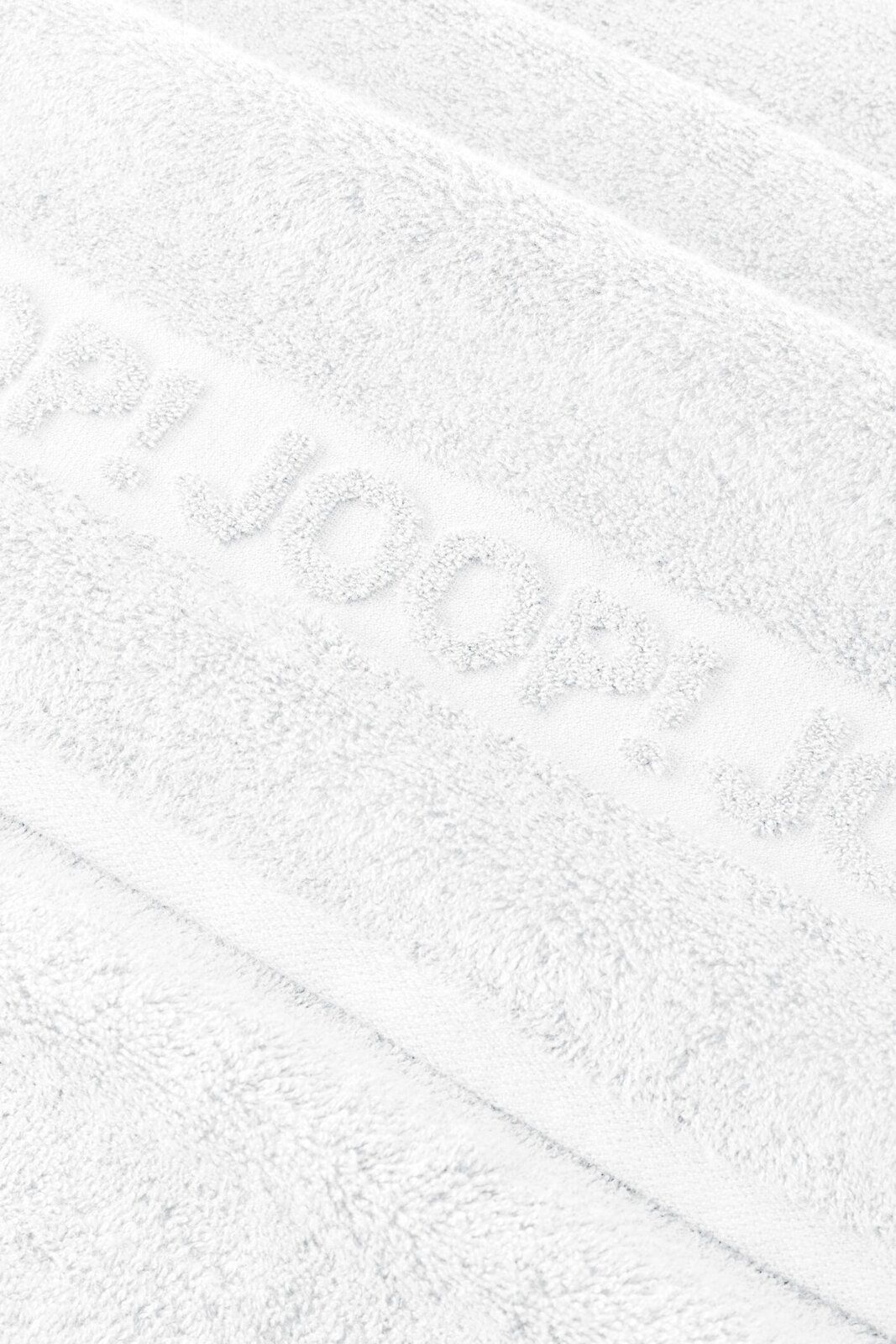 Nahaufnahme in schräger Draufsicht auf ein weißes, flauschiges Handtuch mit eingewebtem Schriftzug „HUGO“.