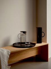 Eine stilvolle, transparente Kanne mit Deckel und Henkel, gefüllt mit Wasser und Eiswürfeln, steht auf einem runden Tablett auf einer Holzbank. Im Hintergrund ist eine schwarze Kanne zu sehen. Die Perspektive ist seitlich aufgenommen.