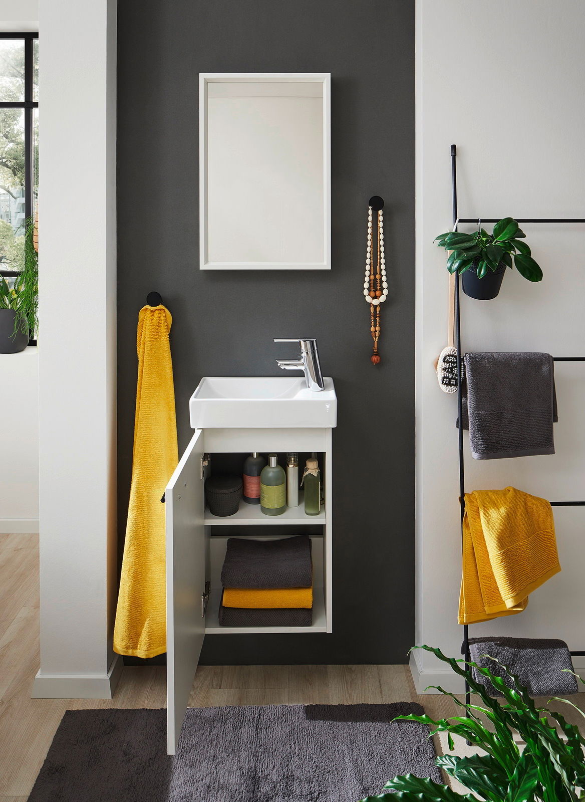 Modernes Gästebad mit weißem Waschbecken und offenem Unterschrank, gelben und grauen Handtüchern, Pflanzen und Accessoires, Frontalansicht.