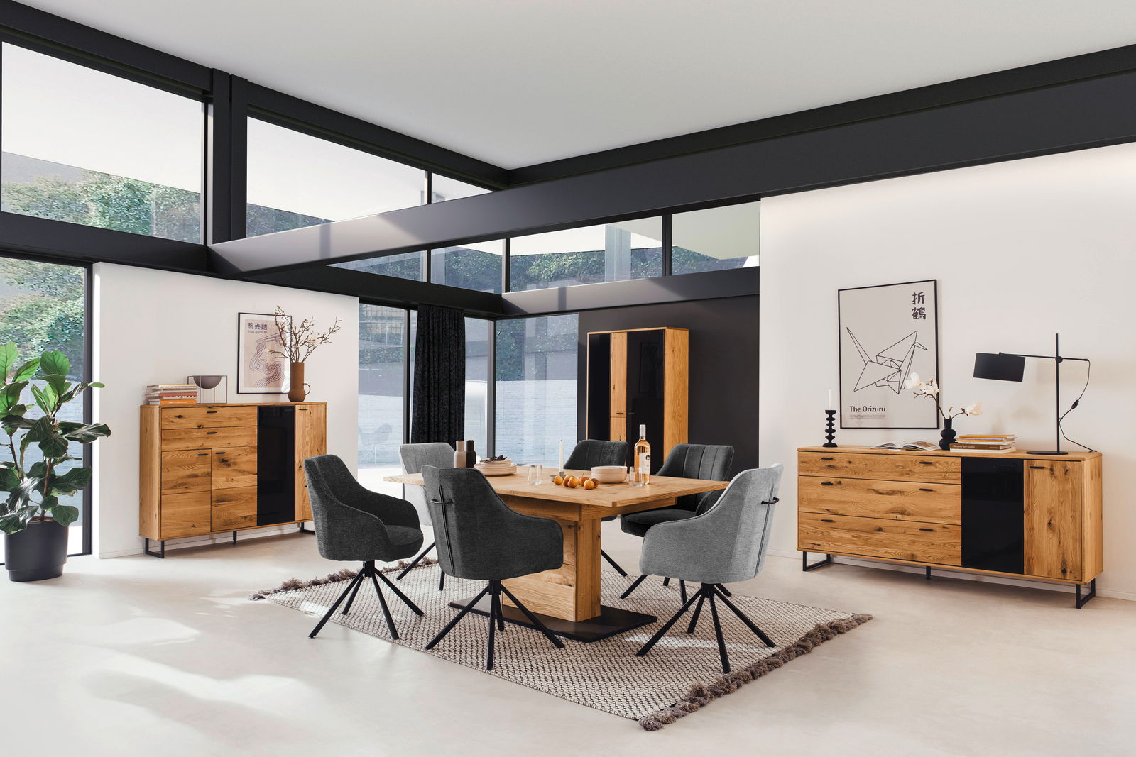 Moderne Esszimmermöbel in einem hellen Raum mit großen Fenstern, bestehend aus einem Holztisch, grauen Stühlen und einem Sideboard, aus der Perspektive des Raumes betrachtet.