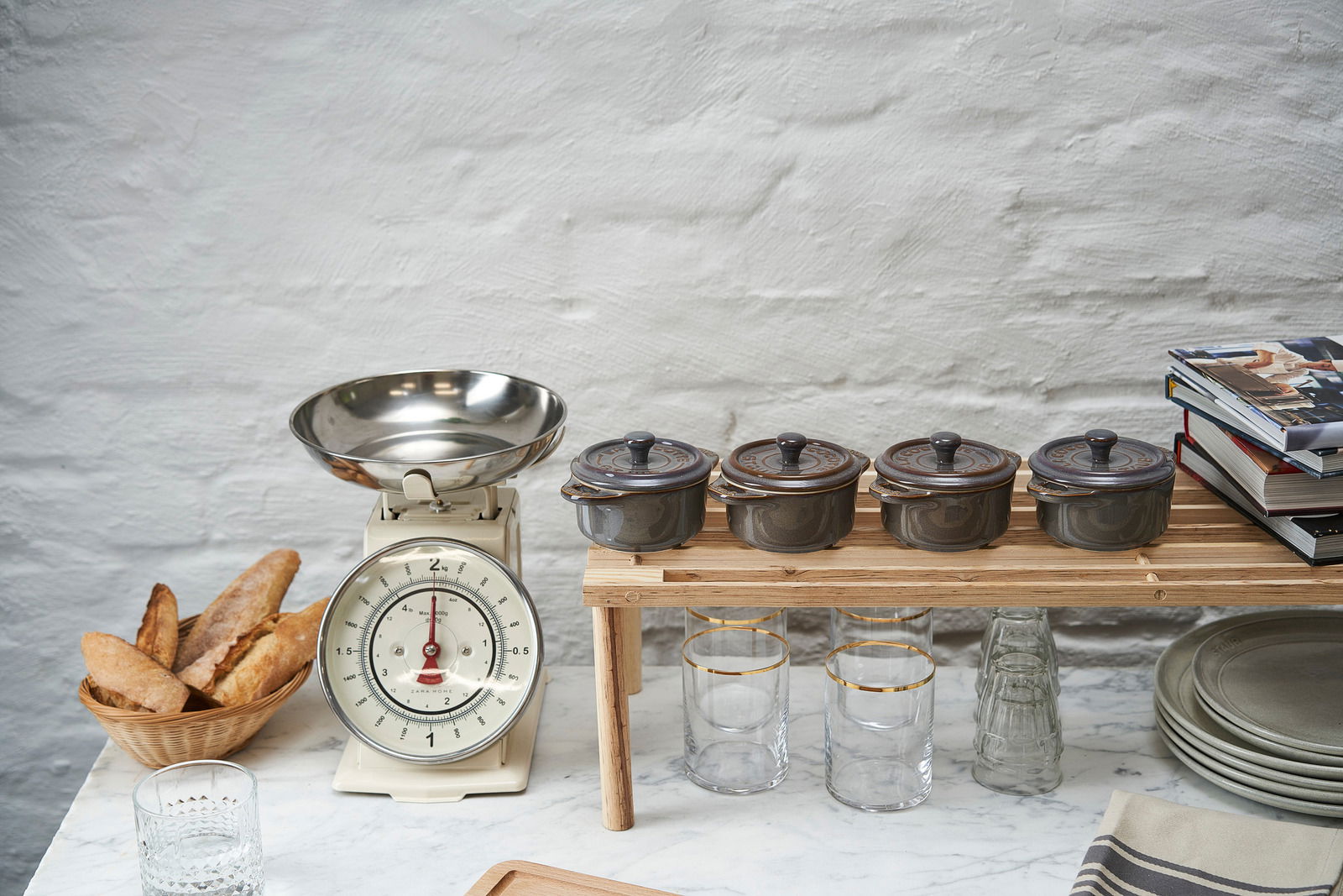 Vier graue Mini-Cocottes auf einem Holzregal, seitliche Perspektive, neben einer Vintage-Küchenwaage und einem Korb mit Baguettes.