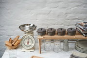 Vier graue Mini-Cocottes auf einem Holzregal, seitliche Perspektive, neben einer Vintage-Küchenwaage und einem Korb mit Baguettes.