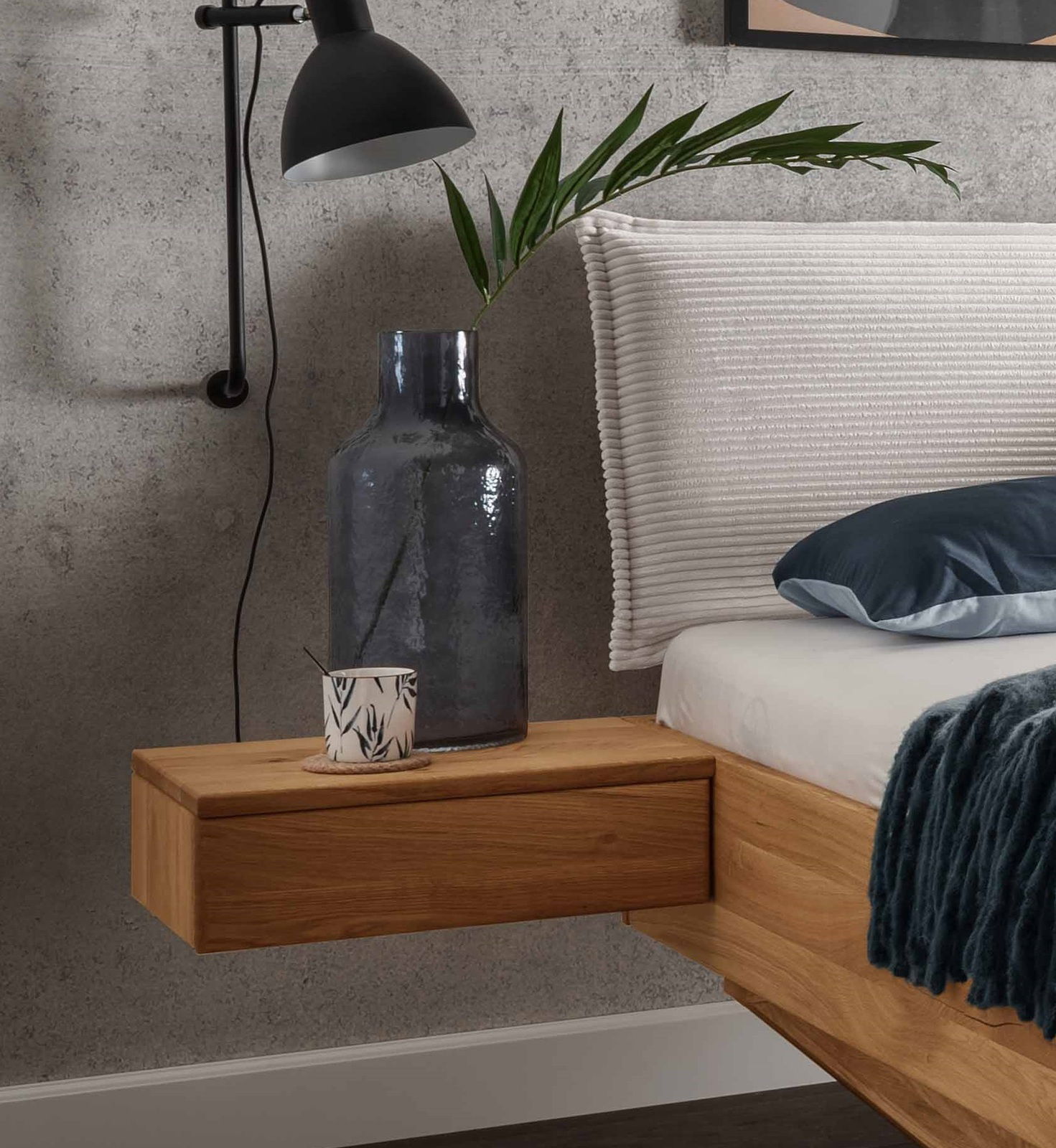 Hängende Nachtkonsole aus Holz, seitliche Perspektive, dekoriert mit einer großen Vase und einer kleinen Tasse, neben einem Bett mit weißem Kopfteil.