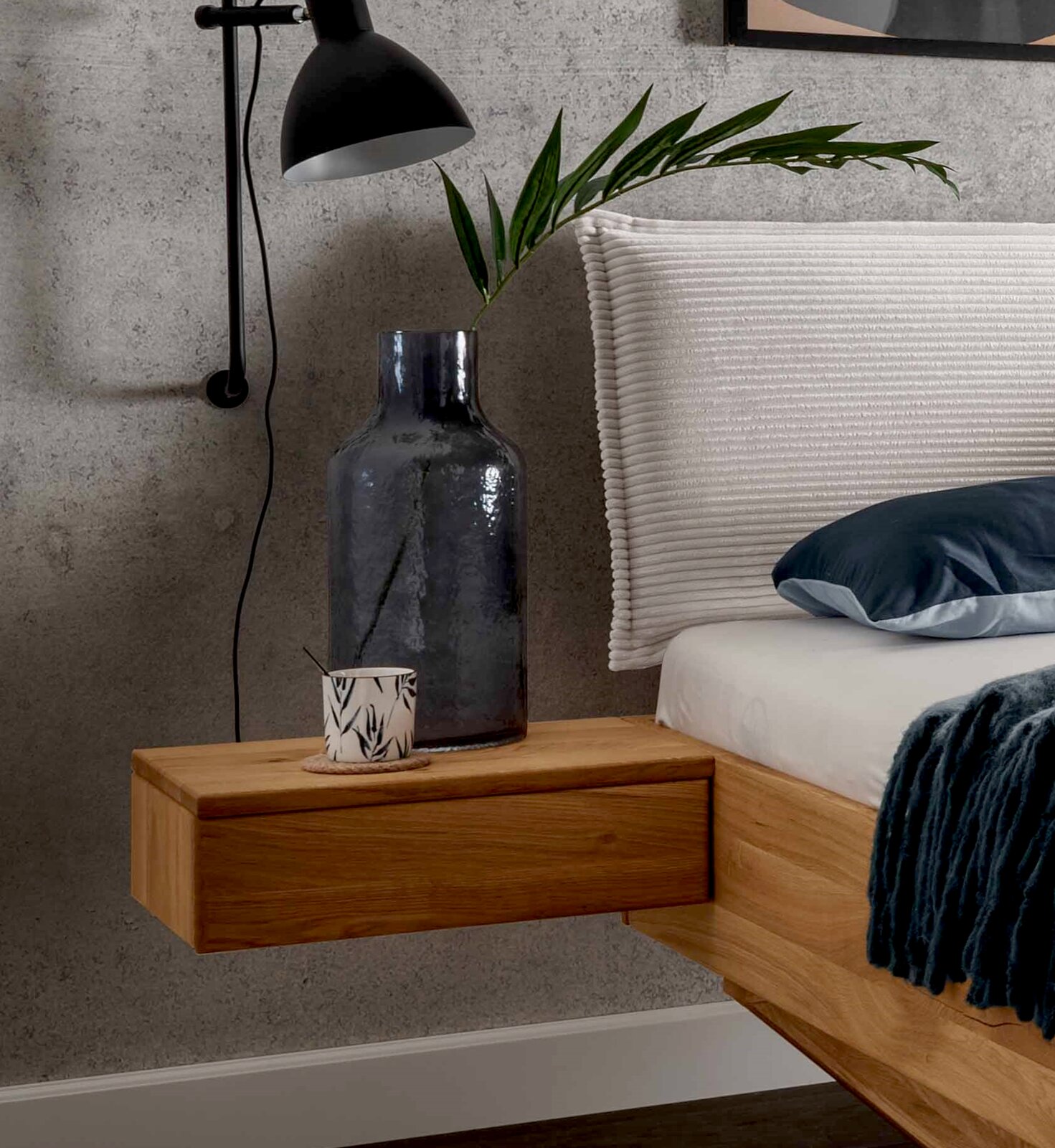 Hängende Nachtkonsole aus Holz, seitliche Perspektive, dekoriert mit einer großen Vase und einer kleinen Tasse, neben einem Bett mit weißem Kopfteil.