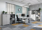 Moderne Büroumgebung mit Schreibtischen, Rollcontainer und Aktenschränken in einem hellen Raum mit großen Fenstern, seitliche Perspektive.
