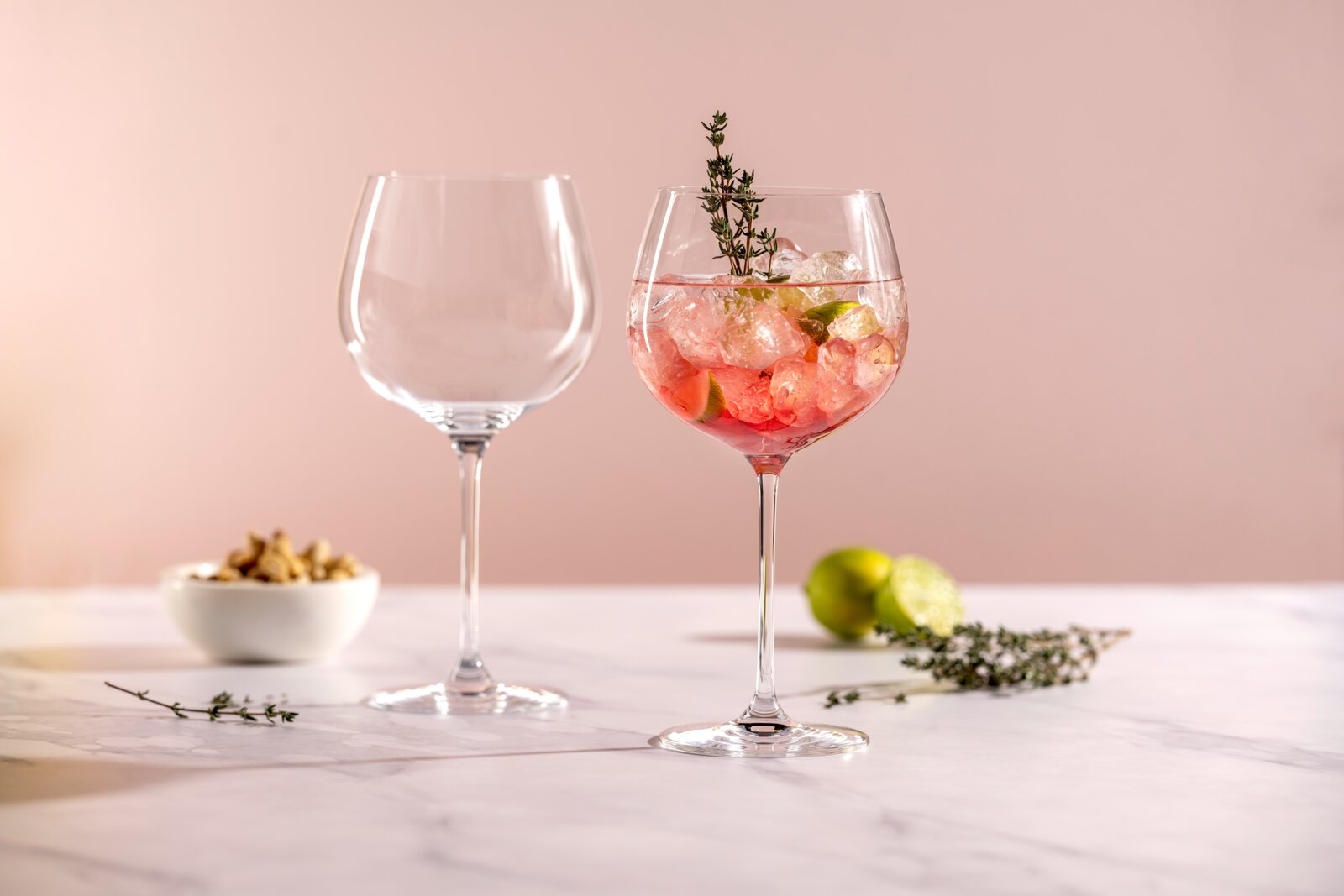 Zwei bauchige Stielgläser auf einer hellen Marmorfläche vor rosafarbenem Hintergrund, rechts ein Glas mit rosafarbenem Cocktail, Eiswürfeln, Limettenscheiben und Thymianzweig, links ein leeres Glas; Perspektive: frontale Tischhöhe.