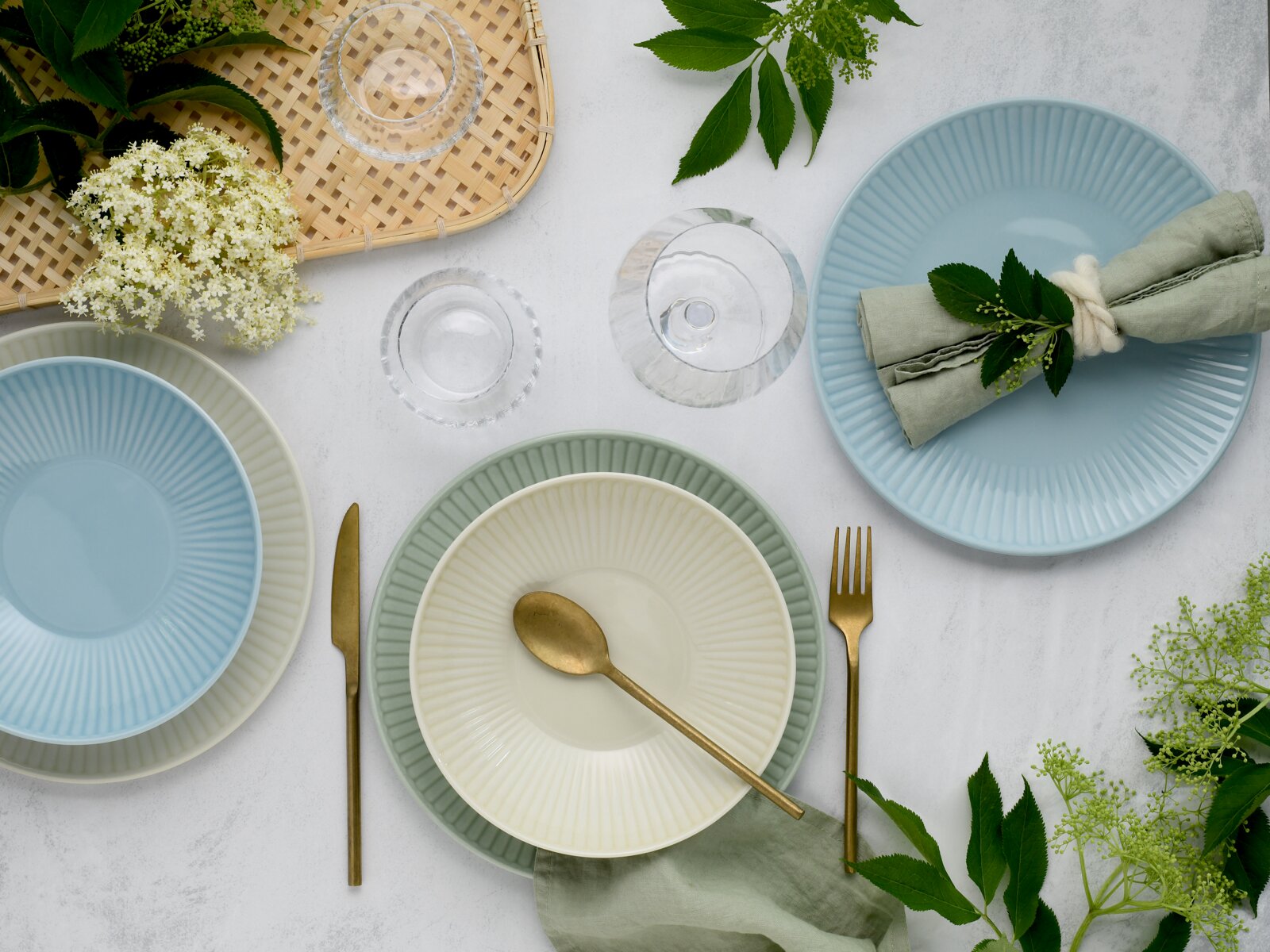 Pastellfarbenes Geschirr- und Tischdeko-Arrangement aus gerippten Tellern und Schalen (blau, grün, creme), goldfarbenem Besteck, Leinenservietten, Glas und Blumen auf hellem Untergrund, fotografiert in Draufsicht von oben.