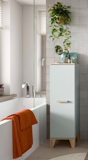 Hochschrank fürs Badezimmer in Hellblau mit Holzgriff und Holzfüßen, seitliche Perspektive im Bad neben Badewanne und Fliesenwand