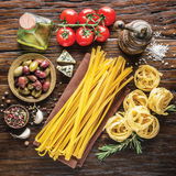 Vogelperspektive auf eine rustikale Holzoberfläche mit frischen Tomaten, Oliven, Knoblauch, Gewürzen, einem Ölfläschchen, einer Pfeffermühle und verschiedenen Pastaformen.