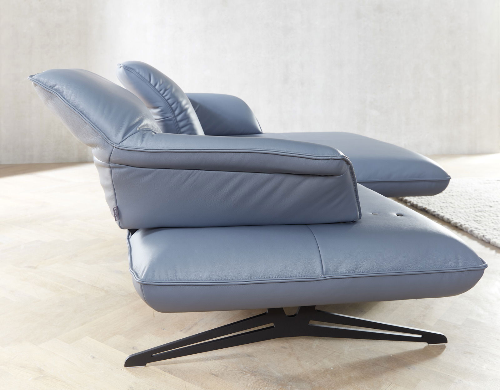 Seitliche Ansicht einer modernen Longchairkombination in blauem Leder mit schwarzem Metallfuß auf hellem Holzboden.