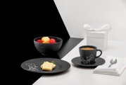 Schwarze Espressotasse mit Untertasse und Mickey-Mouse-Motiv, daneben schwarzer Teller mit Butter, Obstschale und weiße Geschenkbox; Aufnahme aus leicht erhöhter schräger Perspektive (Dreiviertelansicht).