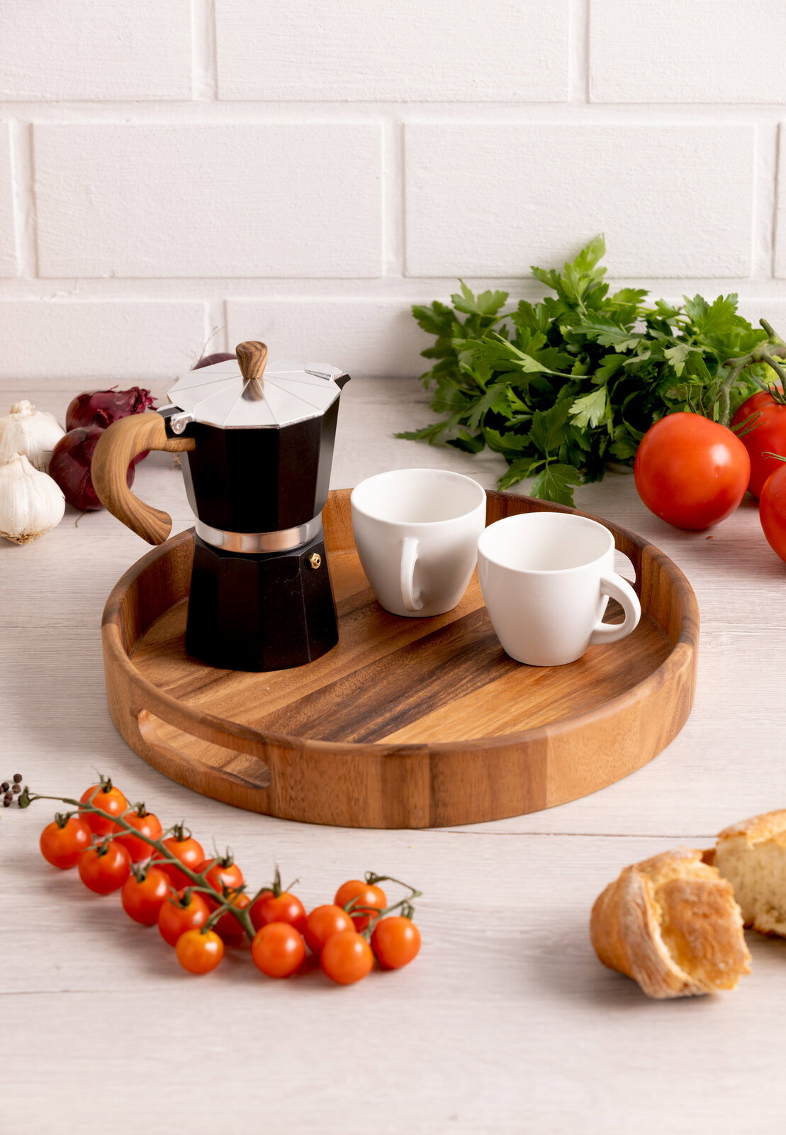 Rundes Holztablett mit zwei weißen Tassen und einer schwarzen Espressokanne, umgeben von frischen Tomaten, Knoblauch und Petersilie, aus der Vogelperspektive.