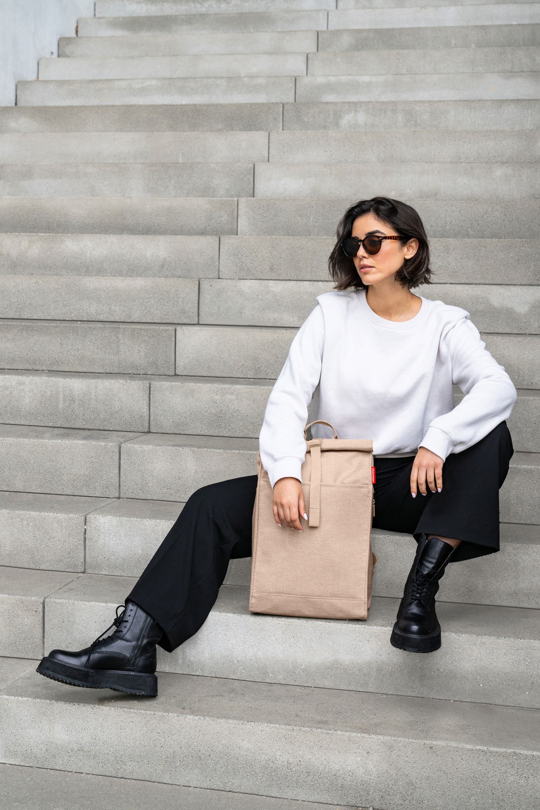 Frau sitzt auf Treppe mit beigem Rolltop-Rucksack, seitliche Perspektive