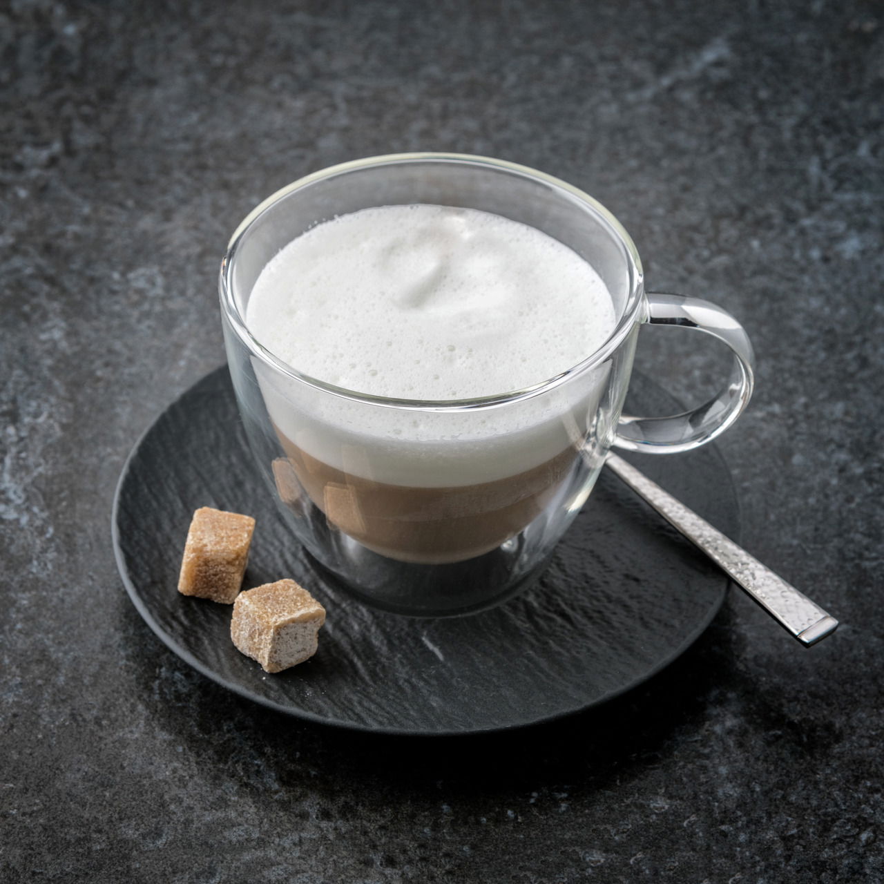 Villeroy & Boch Untertasse MANUFACTURE ROCK Cappuccinountertasse mit Glasbecher, gefüllt mit Cappuccino und Milchschaum, von oben fotografiert, auf einem dunklen Untergrund mit zwei Zuckerwürfeln und einem Löffel.