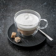 Cappuccinountertasse mit Glasbecher, gefüllt mit Cappuccino und Milchschaum, von oben fotografiert, auf einem dunklen Untergrund mit zwei Zuckerwürfeln und einem Löffel.