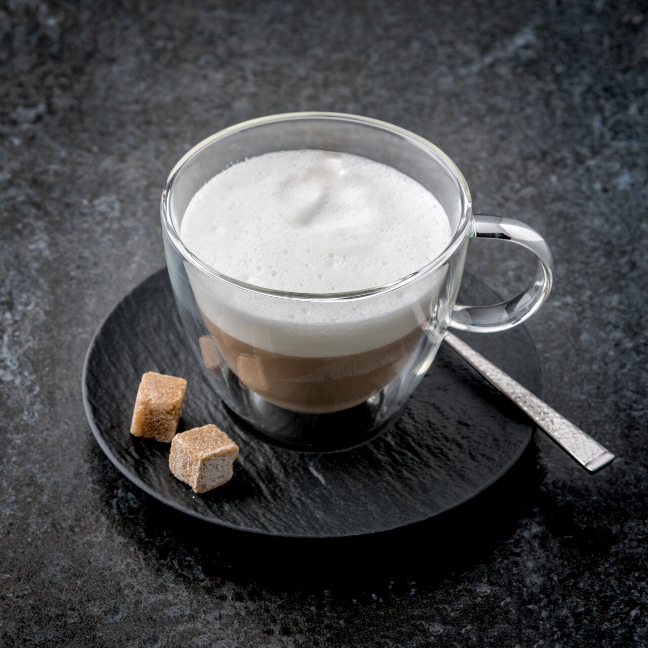 Cappuccinountertasse mit Glasbecher, gefüllt mit Cappuccino und Milchschaum, von oben fotografiert, auf einem dunklen Untergrund mit zwei Zuckerwürfeln und einem Löffel.