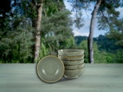 Stapel aus fünf gesprenkelten Keramikschalen in Beige mit braunem Rand, eine Schale vorne liegend, aufgenommen in frontaler Perspektive auf einem Holztisch vor unscharfem Waldhintergrund