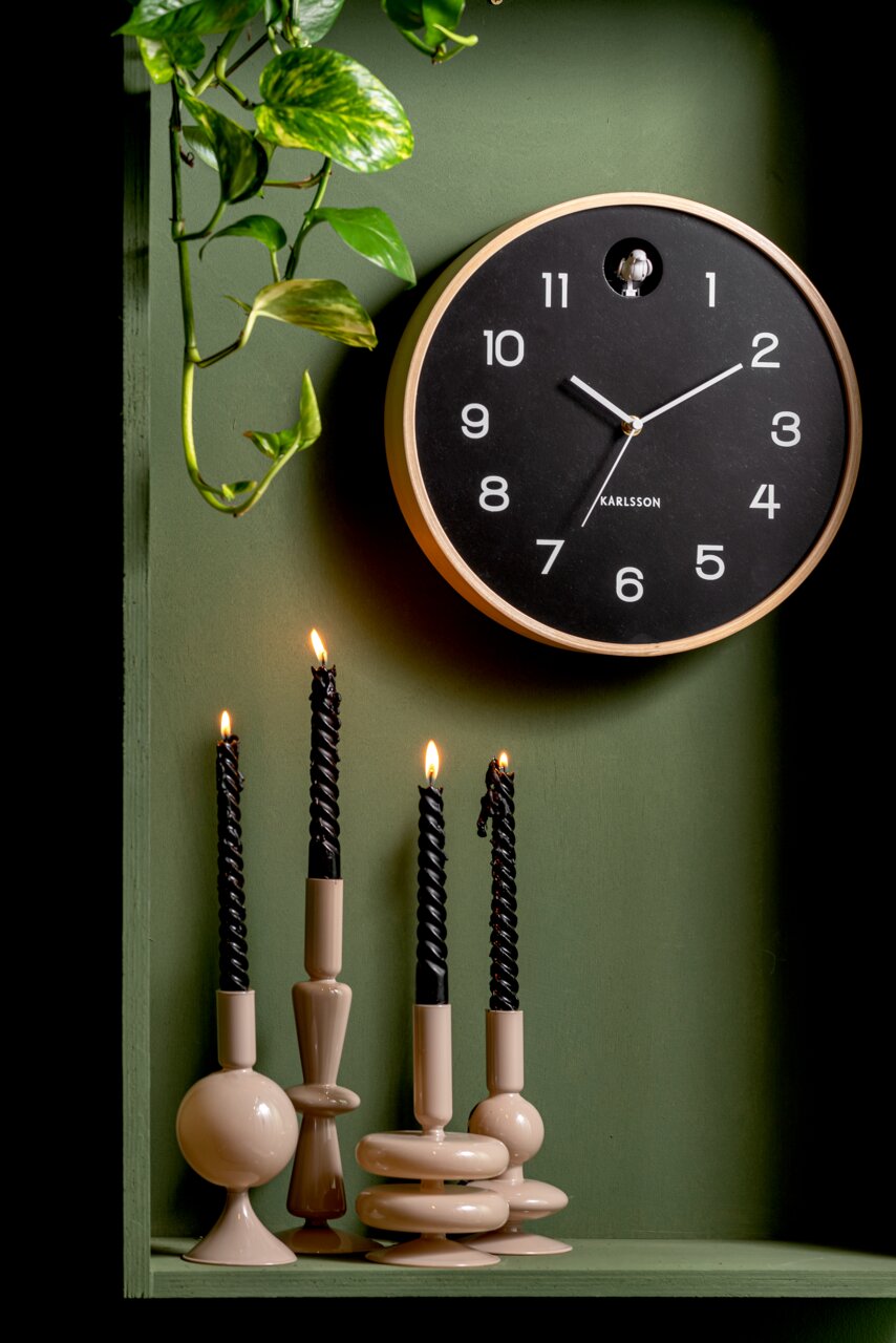 Moderne wandklok met een zwarte wijzerplaat en een witte rand, opgehangen aan een groene muur, omringd door brandende zwarte kaarsen in stijlvolle houders. Vooraanzicht.
