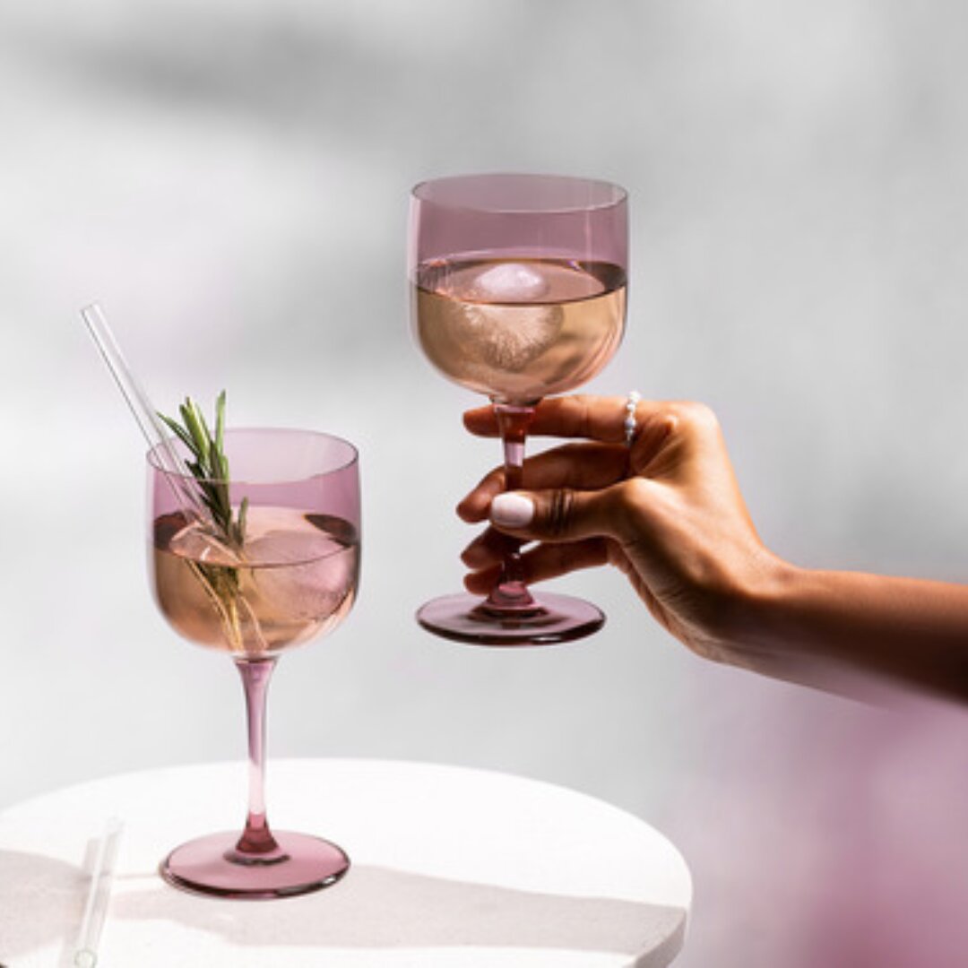 Zwei rosafarbene Weinkelche, einer in der Hand gehalten, der andere auf einem Tisch, mit Eiswürfeln und Rosmarin, seitliche Perspektive.