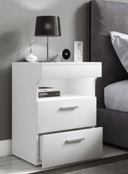 Weiße moderne Nachtkommode mit zwei Schubladen (untere geöffnet) und Ablagefach, daneben Bett; Ansicht schräg von vorn in einem Schlafzimmer mit Lampe, Uhr und Deko auf der Oberfläche