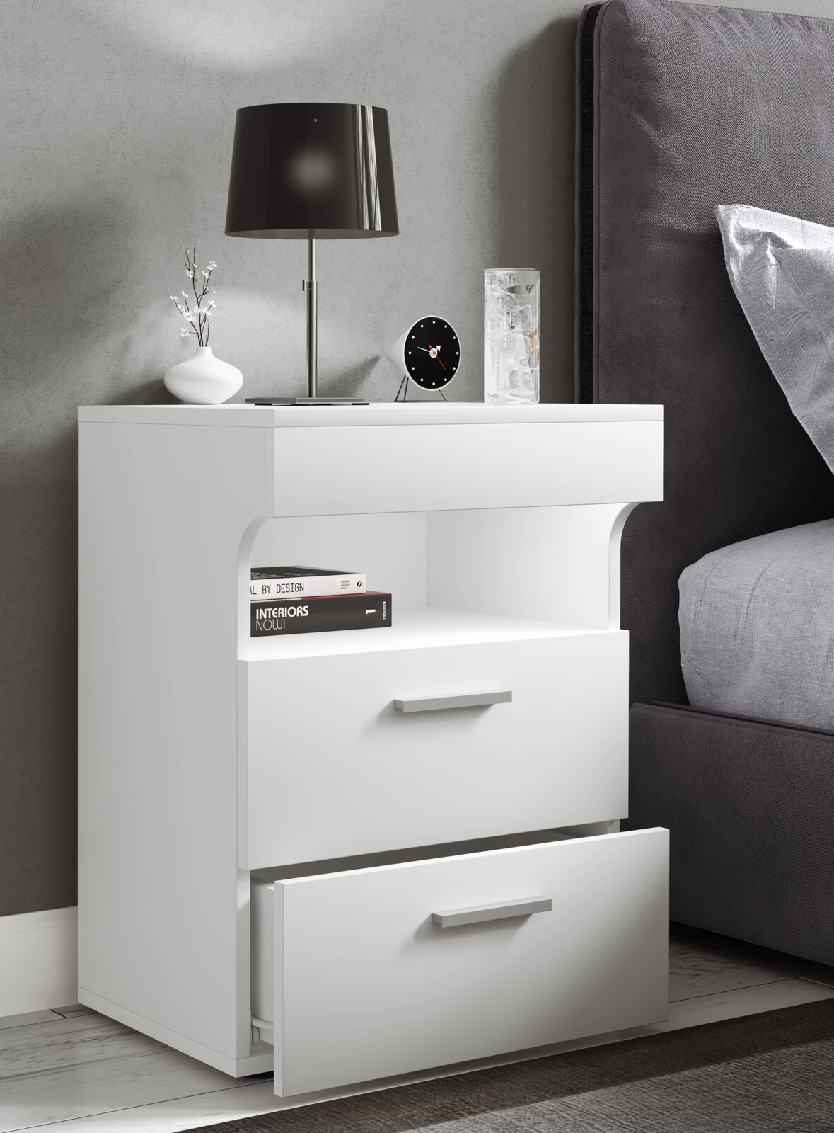 Weiße moderne Nachtkommode mit zwei Schubladen (untere geöffnet) und Ablagefach, daneben Bett; Ansicht schräg von vorn in einem Schlafzimmer mit Lampe, Uhr und Deko auf der Oberfläche