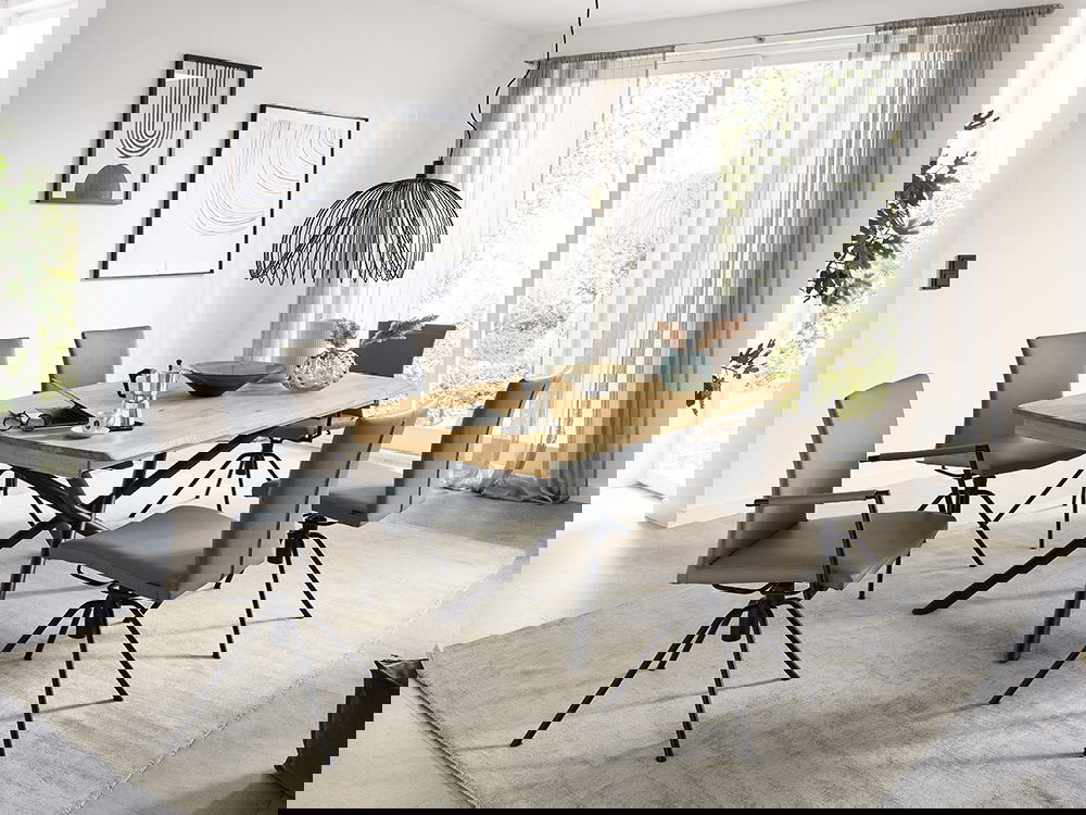 Das Bild zeigt ein modernes Esszimmer. In der Mitte steht ein großer Holztisch mit schwarzen Beinen. Um den Tisch stehen sechs bequeme Stühle mit braunen Polstern. Über dem Tisch hängt eine elegante Lampe aus schwarzem Metall. Helle Vorhänge und große Fen