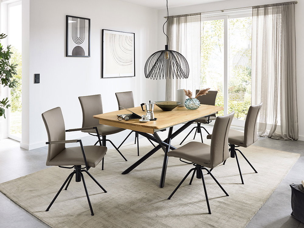 Das Bild zeigt ein modernes Esszimmer. In der Mitte steht ein großer Holztisch mit schwarzen Beinen. Um den Tisch stehen sechs bequeme Stühle mit braunen Polstern. Über dem Tisch hängt eine elegante Lampe aus schwarzem Metall. Helle Vorhänge und große Fen