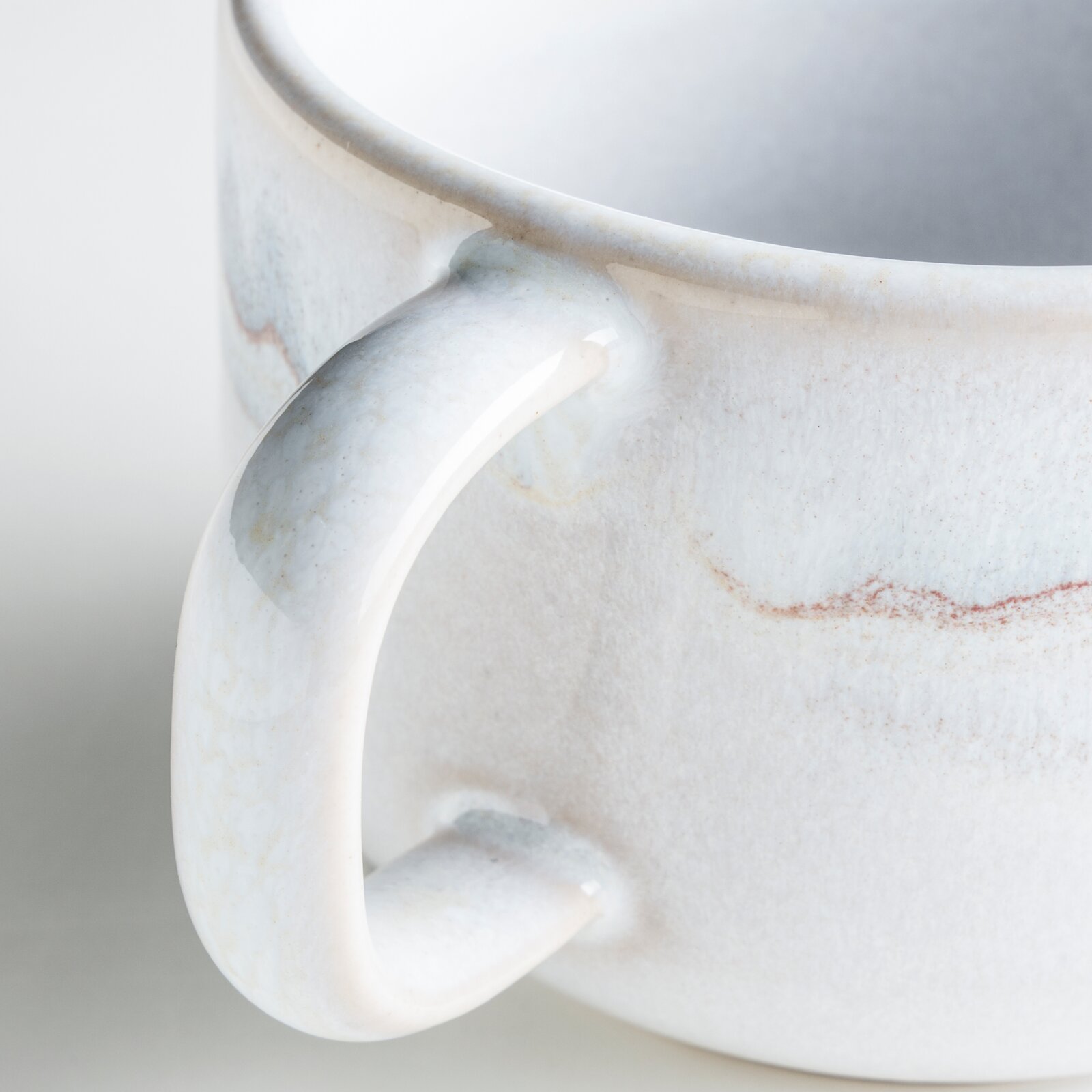 Nahaufnahme einer glasierten Keramik-Tasse in Weiß mit dezenten braun-roten Sprenkeln und sichtbarem Henkel, Perspektive seitlich leicht von oben