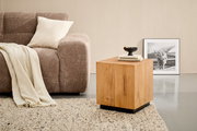 Quadratischer Beistelltisch aus hellem Massivholz mit schwarzem Sockel neben einem braunen Sofa auf Teppich, fotografiert in leichter Seitenansicht von vorn.