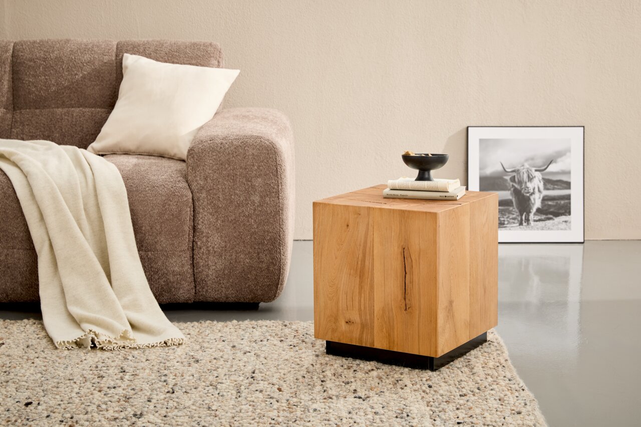 Quadratischer Beistelltisch aus hellem Massivholz mit schwarzem Sockel neben einem braunen Sofa auf Teppich, fotografiert in leichter Seitenansicht von vorn.