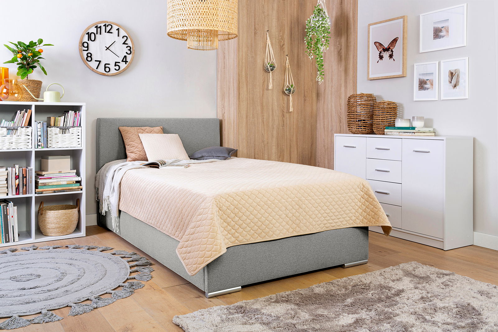 Gemütliches Schlafzimmer mit einem grauen Boxbett, dekoriert mit Kissen und einer beigen Steppdecke, umgeben von modernen Möbeln und Pflanzen. Perspektive von der Seite.