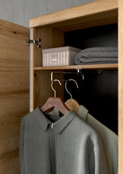 Innenansicht eines Holzschranks mit geöffneter Tür, zeigt Kleiderbügel mit Kleidung, ein Regal mit einer Box und einem gefalteten Handtuch.