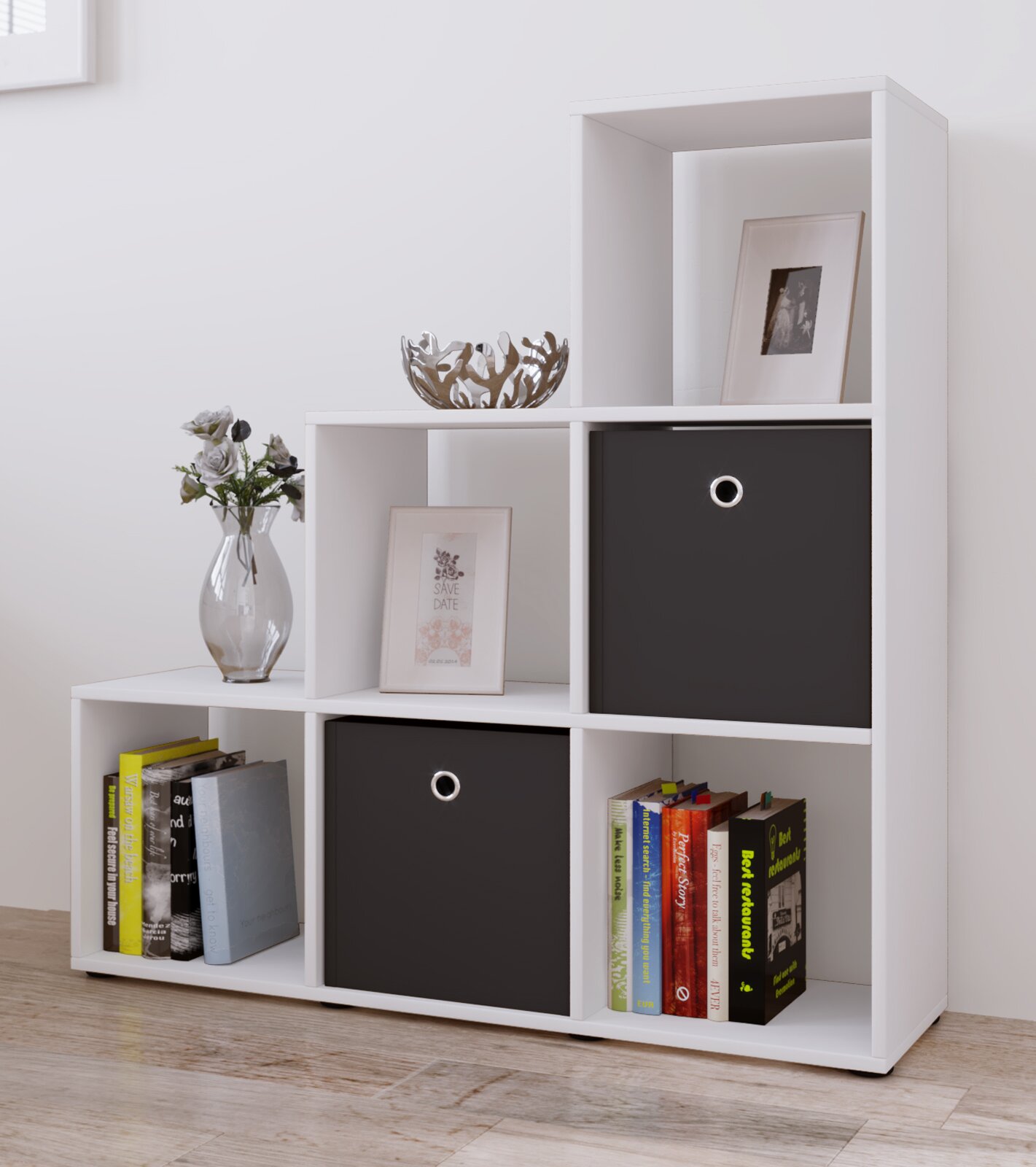 Weißes, modernes Stufenregal mit offenen Fächern und zwei schwarzen Stoffboxen, dekoriert mit Büchern, Vase und Bilderrahmen; Ansicht schräg von vorn (Front-/Seitenperspektive).