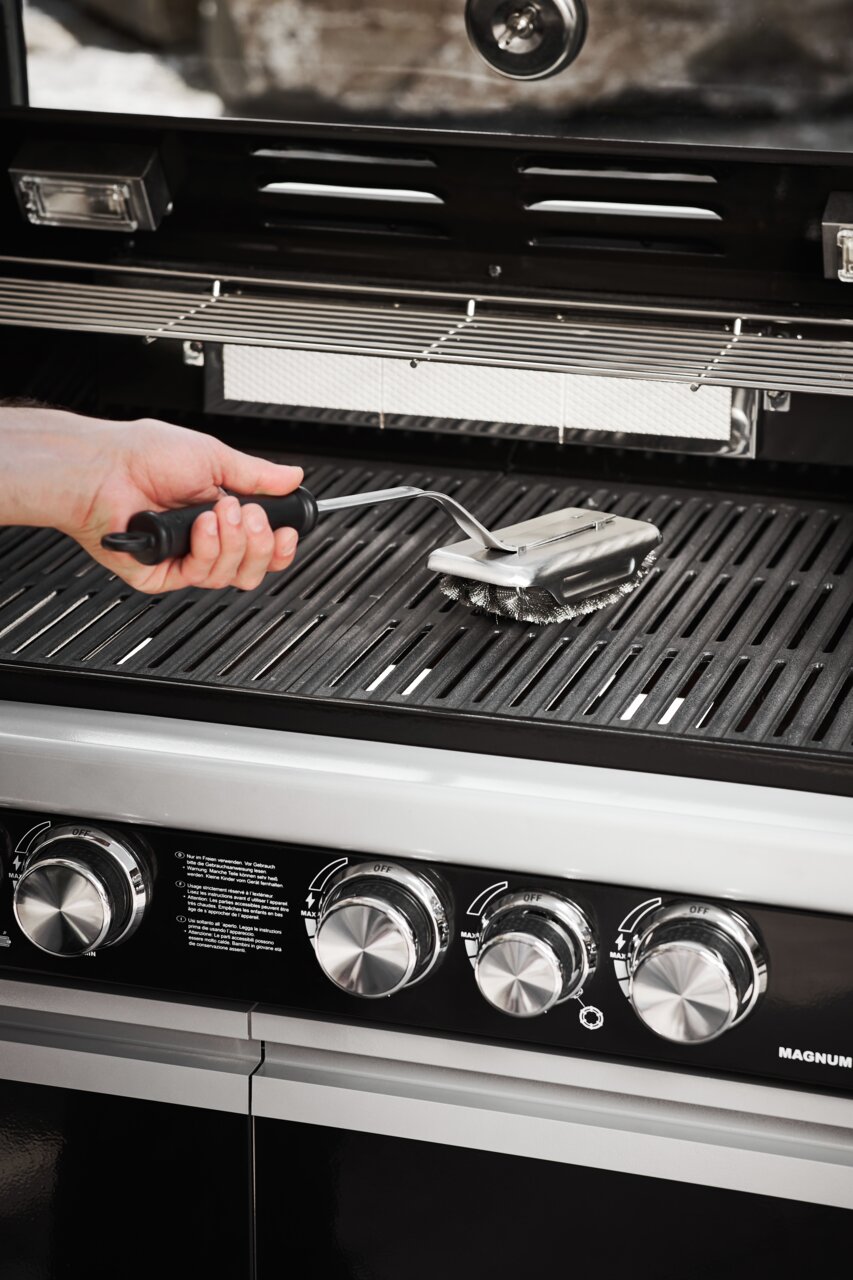 Rösle Reinigungsbürste SlideX BBQ Hand hält eine BBQ Reinigungsbürste SlideX über einem Grillrost, seitliche Perspektive