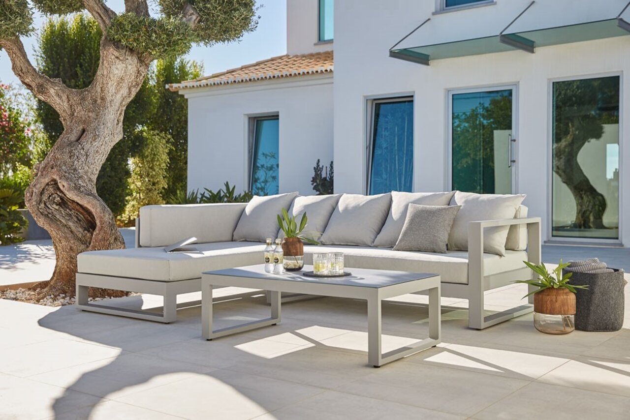 Amatio Loungeset (links) WILLIS Modernes Lounge-Set in hellem Grau auf einer Terrasse, mit einem Baum im Hintergrund und einem Haus mit großen Fenstern. Perspektive von vorne links.