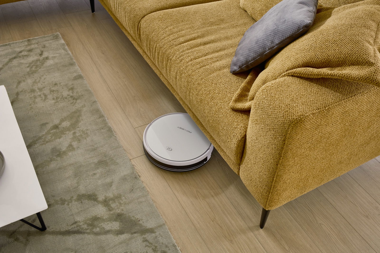 Ein gelbes Ecksofa von Dieter Knoll, Modell MORINO, mit einem grauen Kissen. Ein Saugroboter reinigt den Boden unter dem Sofa. Perspektive von oben.