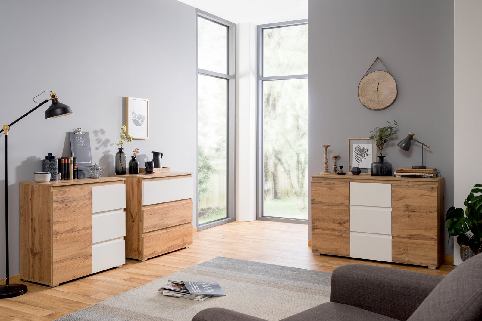 Moderne Kommode aus Holz mit weißen Schubladenfronten, in einem hellen Raum mit großen Fenstern, aus seitlicher Perspektive fotografiert.
