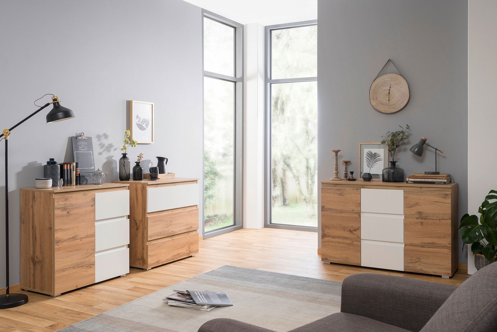 Moderne Kommode aus Holz mit weißen Schubladenfronten, in einem hellen Raum mit großen Fenstern, aus seitlicher Perspektive fotografiert.