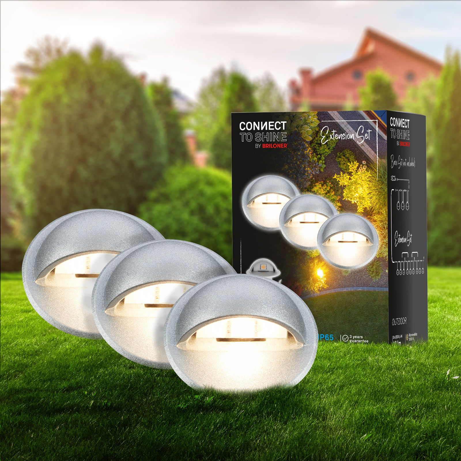 Erweiterungsset aus Edelstahl mit runden, leuchtenden Lampen, präsentiert auf einer grünen Wiese mit Verpackung im Hintergrund. Perspektive: Frontalansicht.