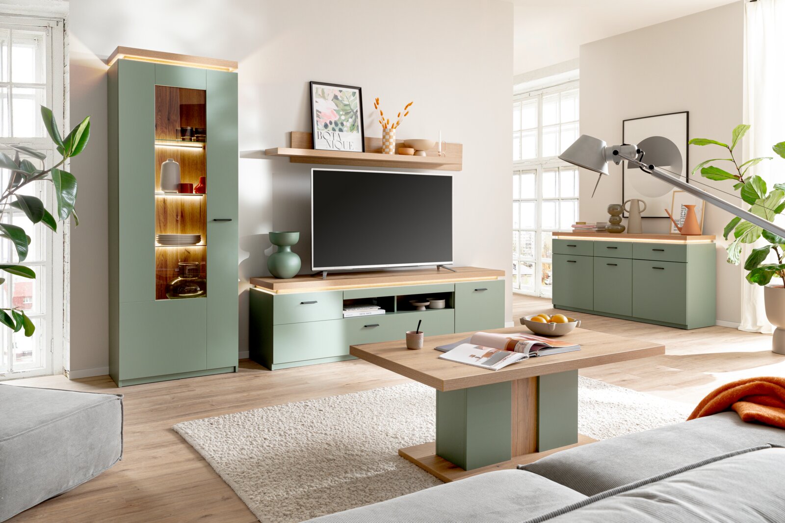 Modernes Wohnzimmer-Set in Salbeigrün mit Holzoptik: Vitrine mit Glasfront und Beleuchtung, TV-Lowboard mit Wandboard, Sideboard und Couchtisch aufgenommen in frontaler Raumansicht mit leichter Perspektive von links.