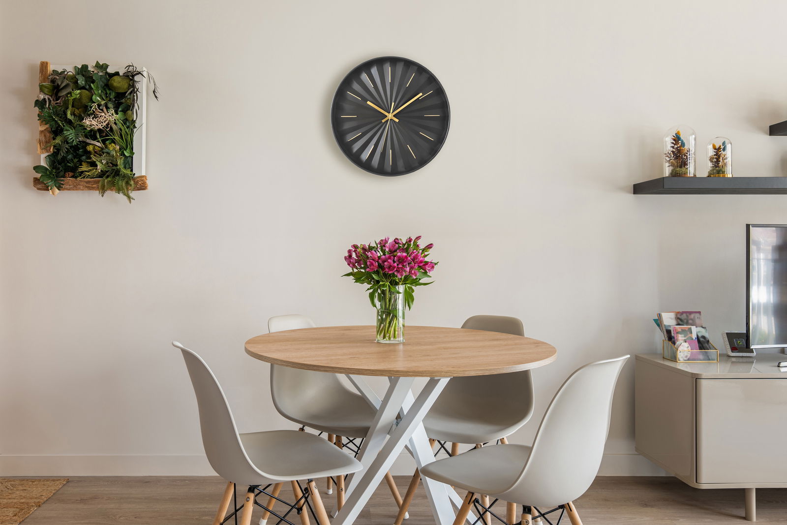 Moderne schwarze Wanduhr mit goldenen Zeigern, zentral über einem Esstisch mit Blumenstrauß, aus frontaler Perspektive.