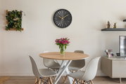 Moderne schwarze Wanduhr mit goldenen Zeigern, zentral über einem Esstisch mit Blumenstrauß, aus frontaler Perspektive.