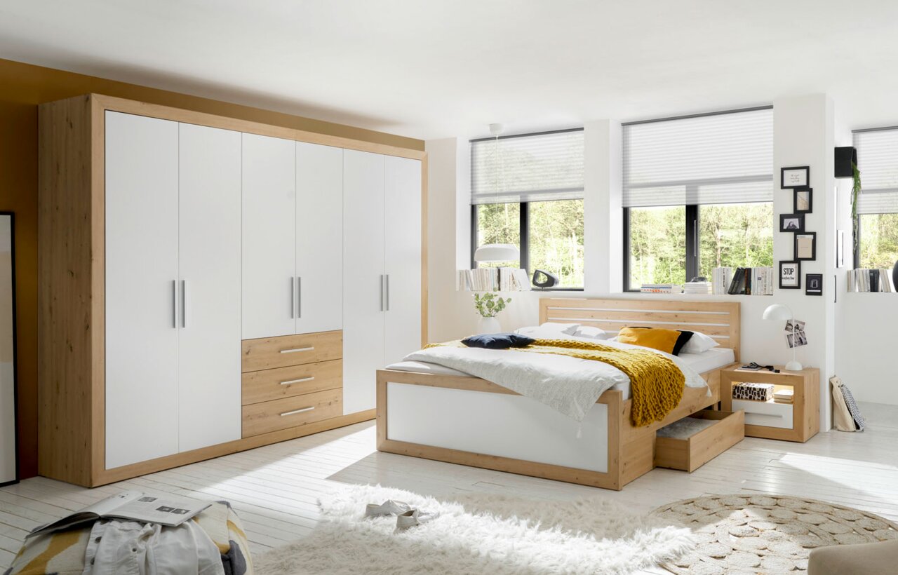 Schlafzimmeransicht mit einem sechstürigen Drehtürenschrank und einer Bettanlage aus Holz und weißen Elementen, seitliche Perspektive.