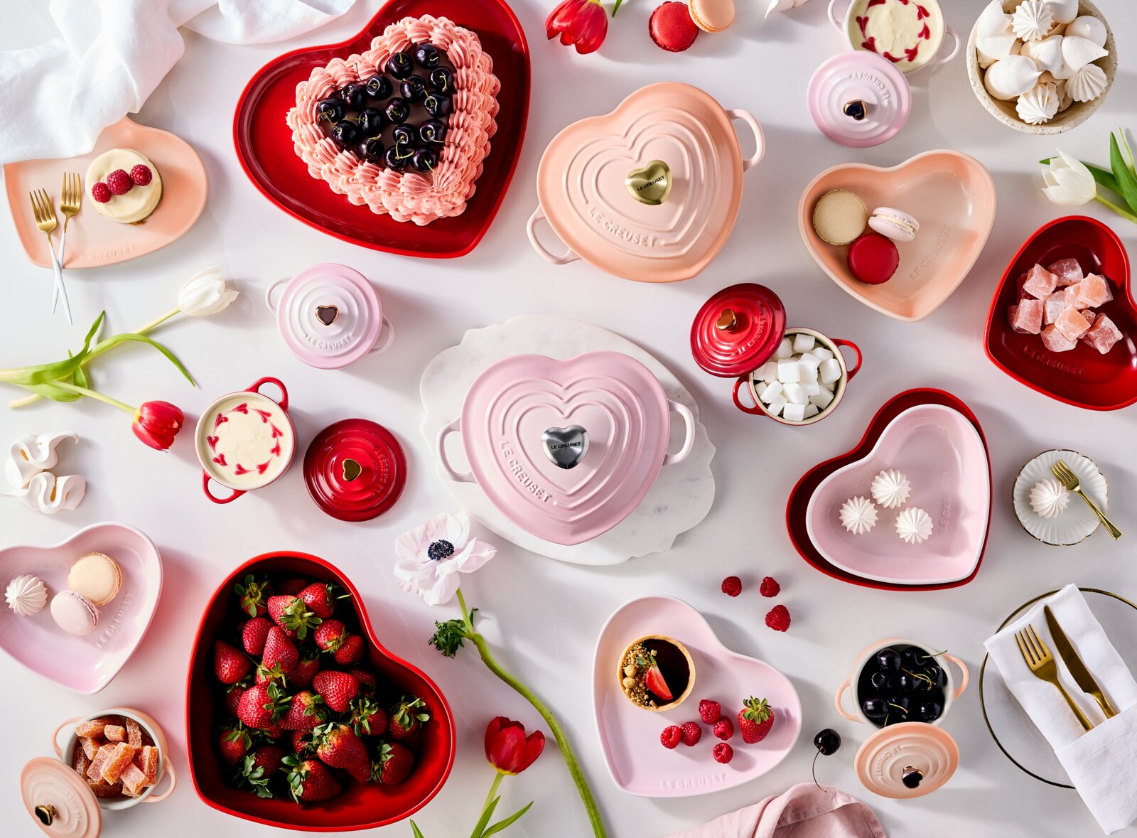 Draufsicht (Flatlay) auf herzförmige Le Creuset Keramikschalen und Mini-Cocottes in Rosa und Rot, dekoriert mit Kuchen, Macarons, Erdbeeren, Kirschen, Marshmallows und Baiser auf hellem Tisch.