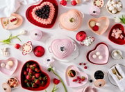 Draufsicht (Flatlay) auf herzförmige Le Creuset Keramikschalen und Mini-Cocottes in Rosa und Rot, dekoriert mit Kuchen, Macarons, Erdbeeren, Kirschen, Marshmallows und Baiser auf hellem Tisch.