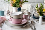 Gestapelte Emaille-Schüsseln in Rosa und Beige auf gedecktem Tisch mit Tellern, Besteck, Gläsern und Frühlingsblumen, fotografiert in leichter Seitenansicht auf Tischhöhe