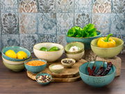 Verschiedene Schalen in einem Buddha Bowl Set, gefüllt mit frischen Zutaten wie Zitronen, Limetten, Salat, Paprika und Gewürzen, auf einem Holztisch vor einer dekorativen Fliesenwand. Perspektive von vorne.