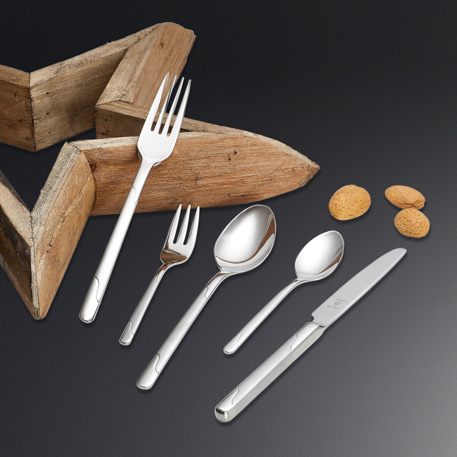 Elegantes Besteckset Viva 60-tlg. aus Edelstahl, bestehend aus Messer, Gabel, Löffel und Teelöffel, auf schwarzem Hintergrund mit Holzstern und Mandeln, aus der Vogelperspektive.