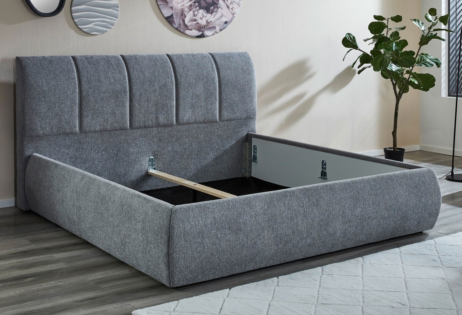 Graues Polsterbettgestell mit hochgeklapptem Lattenrost, seitliche Perspektive, moderne Einrichtung mit Pflanzen und Wanddekoration.