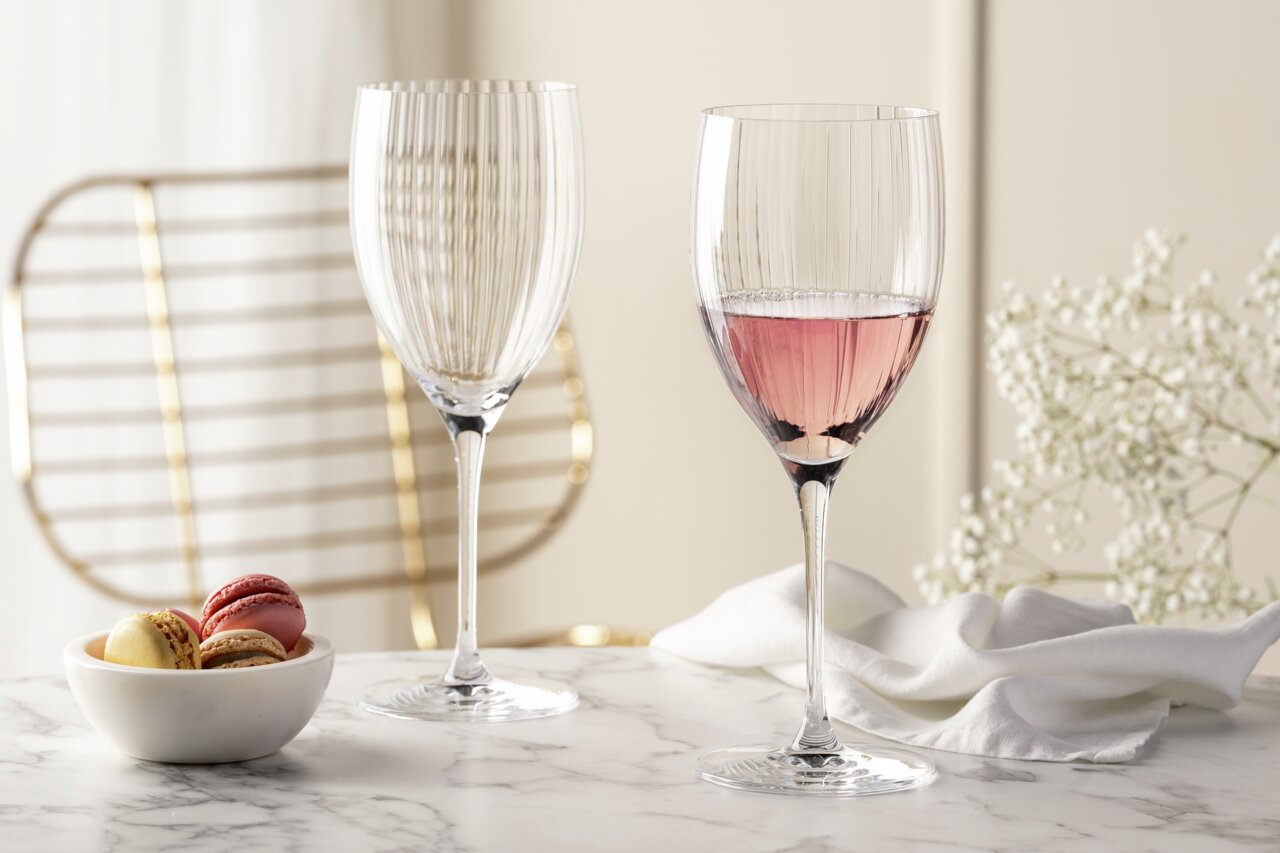 Zwei elegante Rotweingläser POESIA 600ml auf einem Marmortisch, eines gefüllt mit roséfarbenem Wein, im Hintergrund eine goldene Stuhllehne und weiße Blumen, seitliche Perspektive.